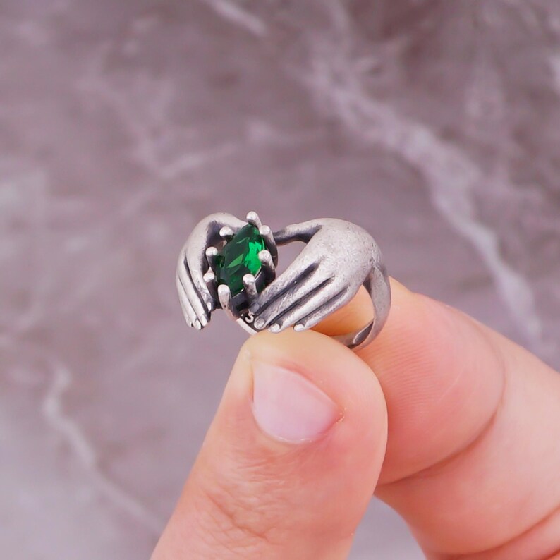 May include: A silver-toned ring featuring a green gemstone held by two hands. The ring has a detailed design, with the hands appearing to cradle the oval-shaped, emerald-colored stone. The ring's band is a simple, rounded design.