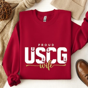 May include: A red sweatshirt with the text "PROUD USCG wife" in white and gold lettering. The letters "USCG" feature anchor designs. The sweatshirt is folded on a white surface, with a tan beanie and brown shoe visible.