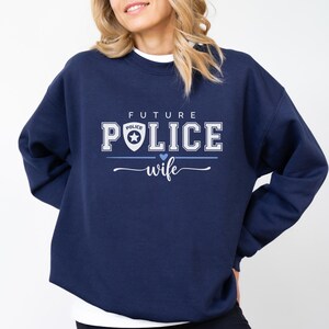 May include: Navy blue sweatshirt with the text "FUTURE POLICE wife" in white and light blue. The word "POLICE" has a police badge graphic. A blue heart is between "POLICE" and "wife".