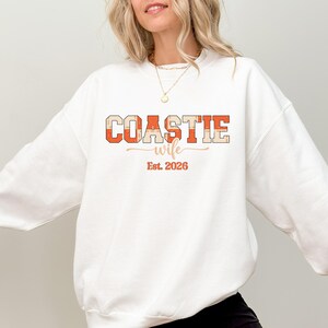May include: White sweatshirt with the text "COASTIE wife Est. 2026" in orange and beige block letters. The word "wife" is in a cursive font. A casual, comfortable garment.