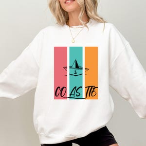 May include: White sweatshirt with a colorful graphic. The design features a boat with an anchor inside a vertical rectangle with pink, teal, and orange stripes. The word "COASTIE" is written in black script below the graphic.