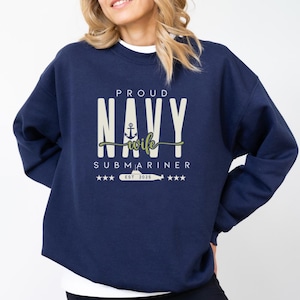 May include: Navy blue sweatshirt with the text "PROUD NAVY wife SUBMARINER" in white and green. The design includes an anchor, stars, and a submarine, with "EST 2025" below. A casual, comfortable garment.