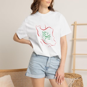 Op de afbeelding: Wit T-shirt met een rode omtrek van een landkaart en groene en rode kalligrafie. Het T-shirt heeft een ronde hals en wordt gedragen met een spijkerbroekshort. Het ontwerp is gecentreerd op de voorkant van het T-shirt.
