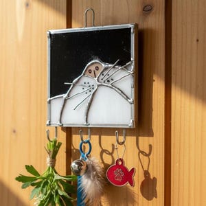 Puede incluir: Llavero con diseño de gato en vidriera, con un diseño de cara de gato en blanco y negro. Incluye tres ganchos con accesorios: un manojo de hierbas, una campana con una pluma y una etiqueta roja con forma de pez. Montado sobre una superficie de madera.