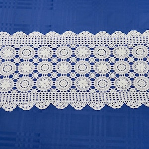 May include: An off-white, rectangular crocheted table runner with a floral design. The runner has a scalloped edge and is displayed on a blue tablecloth. The intricate design features repeating circular motifs.