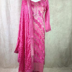 May include: A pink and gold embroidered kurta with a matching dupatta. The kurta has a V-neck and long sleeves. The dupatta is a sheer fabric with a floral pattern.