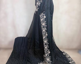 May include: A black saree with intricate floral embroidery in a contrasting light color. The saree is draped over a mannequin, showcasing the detailed design along the borders and scattered across the fabric. Small, sparkling embellishments add a touch of glamour.