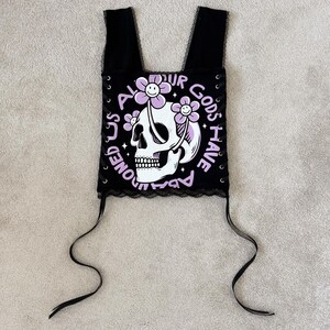 Puede incluir: Top corto negro con un gr&aacute;fico de calavera y el texto "ALL OUR GODS HAVE ABANDONED US". La calavera est&aacute; adornada con flores moradas sonrientes. El top tiene tirantes negros y ribete de encaje.