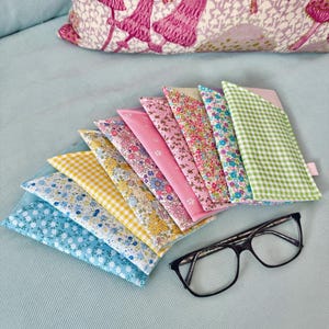 Può includere: Un set di dieci custodie per occhiali in tessuto colorato con motivi floreali e a quadri. Le custodie sono disposte a ventaglio su una superficie blu chiaro. Un paio di occhiali neri è appoggiato sulla superficie.