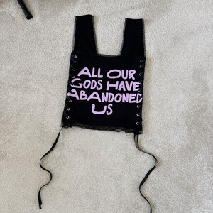 Puede incluir: Top negro con el texto "ALL OUR GODS HAVE ABANDONED US" en lavanda. El top tiene un ribete de encaje y detalles de ojal con lazos de cinta negra. El top tiene dos tirantes.