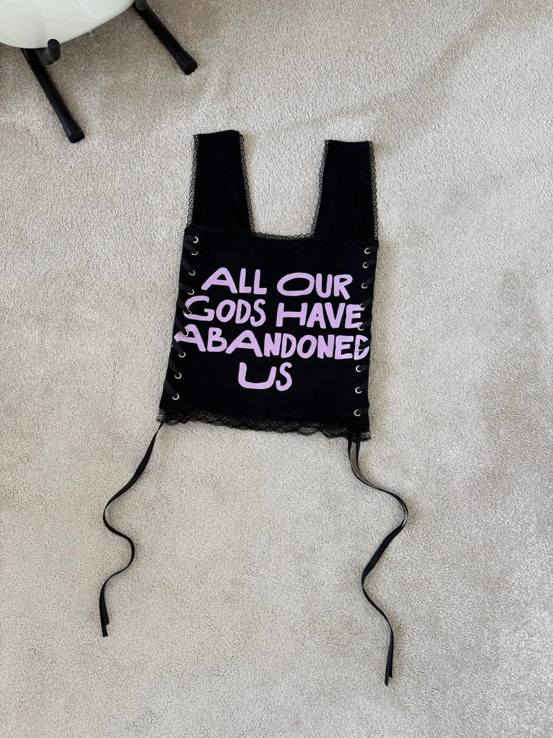 Puede incluir: Top negro con el texto "ALL OUR GODS HAVE ABANDONED US" en lavanda. El top tiene un ribete de encaje y detalles de ojal con lazos de cinta negra. El top tiene dos tirantes.