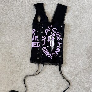 Puede incluir: Top de cors&eacute; negro con ribete de encaje y lazos de cinta. La parte superior presenta el texto "ALL GODS HAVE BONES" en lavanda, junto con un gr&aacute;fico floral. El cors&eacute; tiene un dise&ntilde;o de cordones en los laterales.
