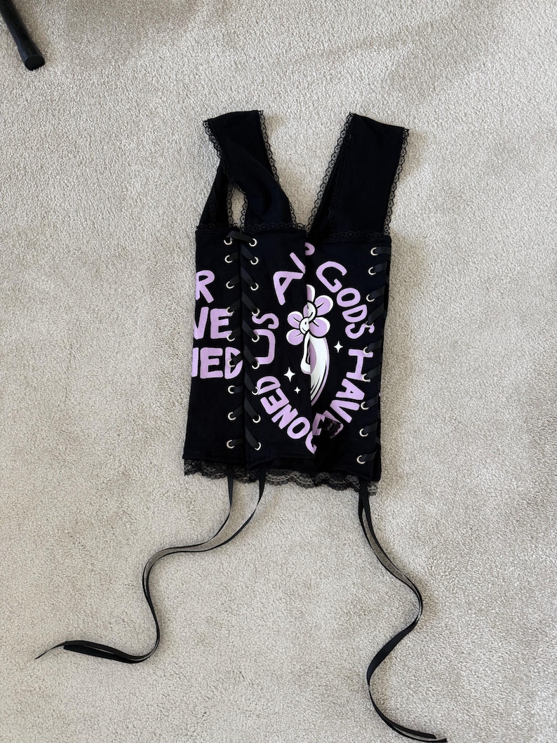 Puede incluir: Top de cors&eacute; negro con ribete de encaje y lazos de cinta. La parte superior presenta el texto "ALL GODS HAVE BONES" en lavanda, junto con un gr&aacute;fico floral. El cors&eacute; tiene un dise&ntilde;o de cordones en los laterales.