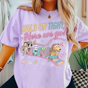 May include: A light purple t-shirt with the text "HOLD ON TIGHT, Here we go!" and a colorful graphic of cartoon characters on a roller coaster. The graphic includes Mickey Mouse, a Stormtrooper, and a dachshund.