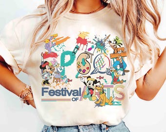 May include: Cream-coloured t-shirt with a vibrant graphic of Disney characters involved in art. The text "Festival of Arts" is displayed in a rainbow design. Mickey Mouse, Donald Duck, and Goofy are featured in the artwork.