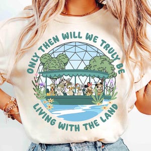 May include: Cream-colored t-shirt featuring a circular graphic with the text "Only Then Will We Truly Be Living With The Land." The graphic depicts a boat with cartoon characters, trees, and water. The shirt has short sleeves.