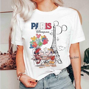 May include: White t-shirt with a graphic of Mickey and Minnie Mouse in Paris. The design includes the Eiffel Tower, balloons, and the text "PARIS Disneyland" in red, white, and blue.