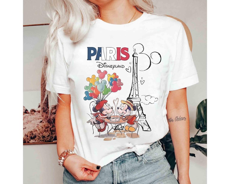 May include: White t-shirt with a graphic of Mickey and Minnie Mouse in Paris. The design includes the Eiffel Tower, balloons, and the text "PARIS Disneyland" in red, white, and blue.