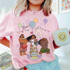 Puede incluir: Camiseta rosa con un gr&aacute;fico de dibujos animados de personajes de una popular pel&iacute;cula animada, globos, una tarta de cumplea&ntilde;os y el n&uacute;mero 5. La palabra "Name" est&aacute; escrita encima de los personajes. Una taza blanca con una carita sonriente se sostiene en la mano.