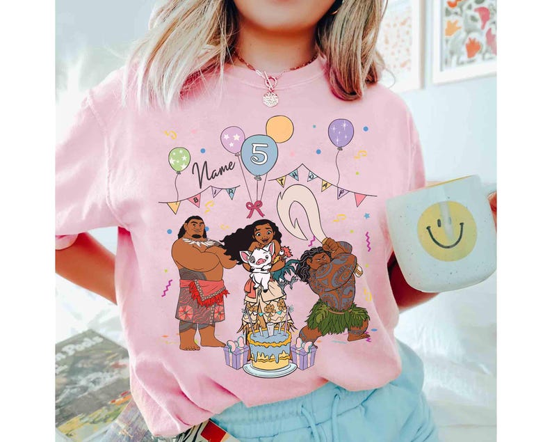 Puede incluir: Camiseta rosa con un gr&aacute;fico de dibujos animados de personajes de una popular pel&iacute;cula animada, globos, una tarta de cumplea&ntilde;os y el n&uacute;mero 5. La palabra "Name" est&aacute; escrita encima de los personajes. Una taza blanca con una carita sonriente se sostiene en la mano.