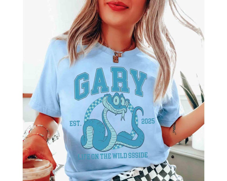 May include: Light blue t-shirt with the name "GARY" in large, block letters. Below the name is a cartoon snake illustration. The text "EST. 2025" and "LIFE ON THE WILD SSSIDE" are also printed on the shirt.