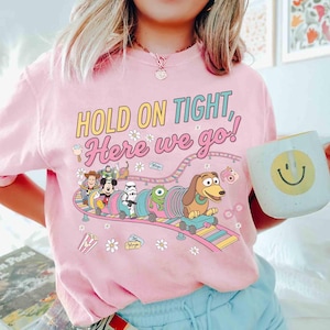 May include: Pink t-shirt with the text "HOLD ON TIGHT, Here we go!" and a colorful graphic of Disney characters on a roller coaster. The shirt also features small flower and popcorn graphics.
