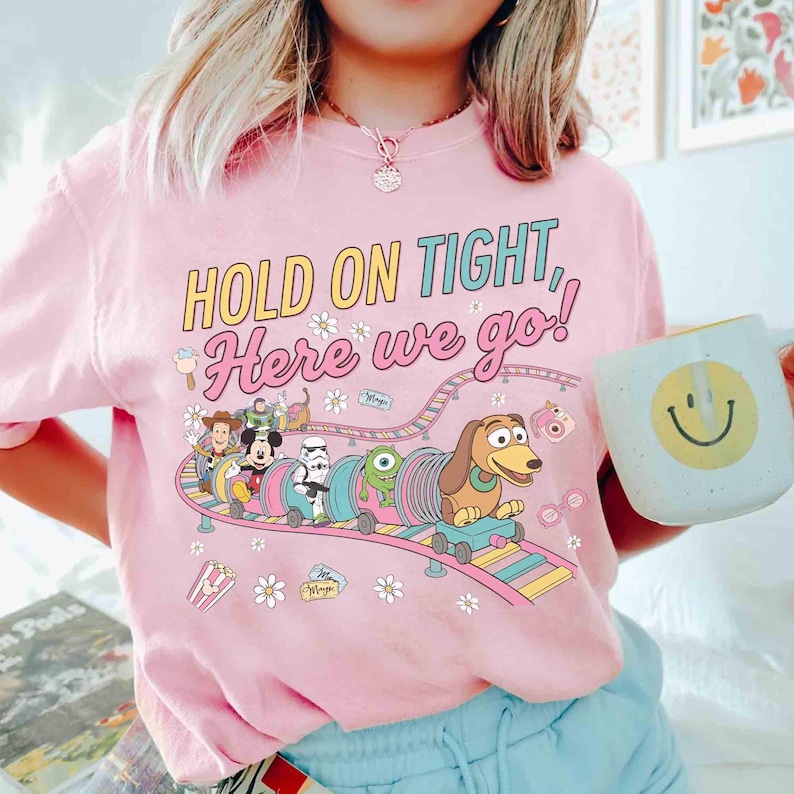May include: Pink t-shirt with the text "HOLD ON TIGHT, Here we go!" and a colorful graphic of Disney characters on a roller coaster. The shirt also features small flower and popcorn graphics.