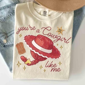 以下が含まれることがあります： クリーム色のTシャツで、赤いカウガールハット、赤い三つ編みの髪型、茶色のカウボーイブーツが描かれています。赤い筆記体で「you're a Cowgirl like me」と書かれ、右上には「INFINITY AND BEYOND」と記された金色の星のバッジがあります。