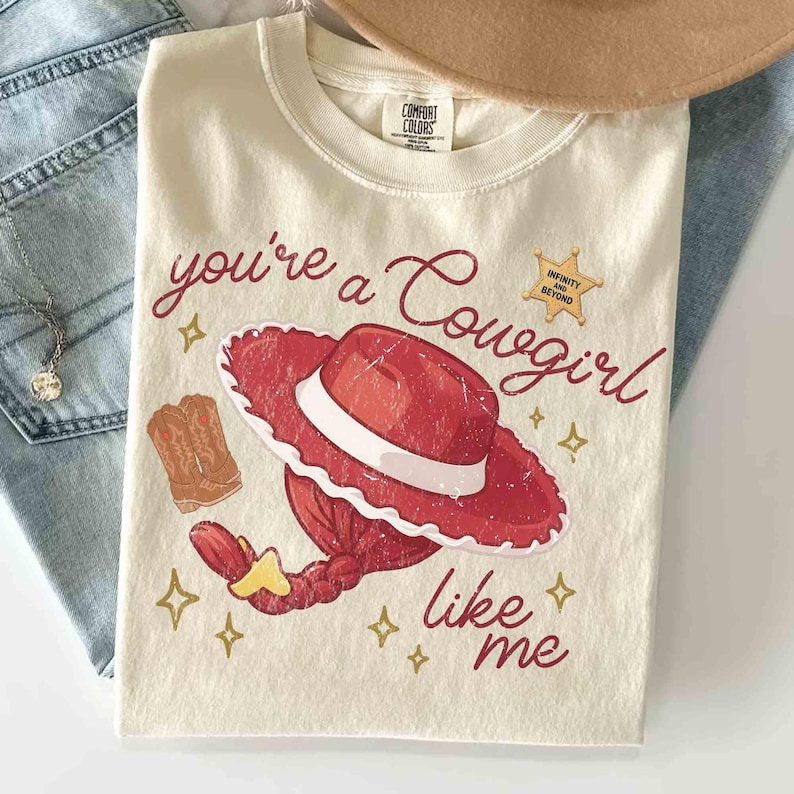 以下が含まれることがあります： クリーム色のTシャツで、赤いカウガールハット、赤い三つ編みの髪型、茶色のカウボーイブーツが描かれています。赤い筆記体で「you're a Cowgirl like me」と書かれ、右上には「INFINITY AND BEYOND」と記された金色の星のバッジがあります。