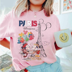 May include: Pink t-shirt with a graphic featuring Mickey and Minnie Mouse in Paris, France. The design includes the Eiffel Tower, balloons, and the text "PARIS Disneyland". A person is wearing the shirt and holding a mug.
