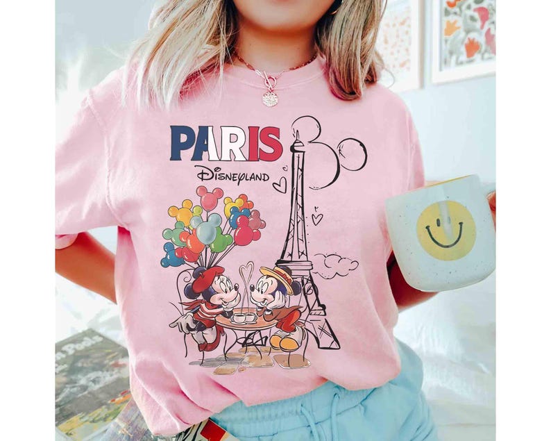 May include: Pink t-shirt with a graphic featuring Mickey and Minnie Mouse in Paris, France. The design includes the Eiffel Tower, balloons, and the text "PARIS Disneyland". A person is wearing the shirt and holding a mug.