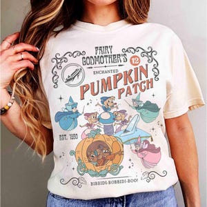 May include: Cream-colored t-shirt featuring a whimsical graphic with the text "Fairy Godmother's Enchanted Pumpkin Patch." The design includes illustrations of Cinderella's mice, fairies, and a pumpkin carriage. The text "Bibbidi-Bobbidi-Boo!" is also included.