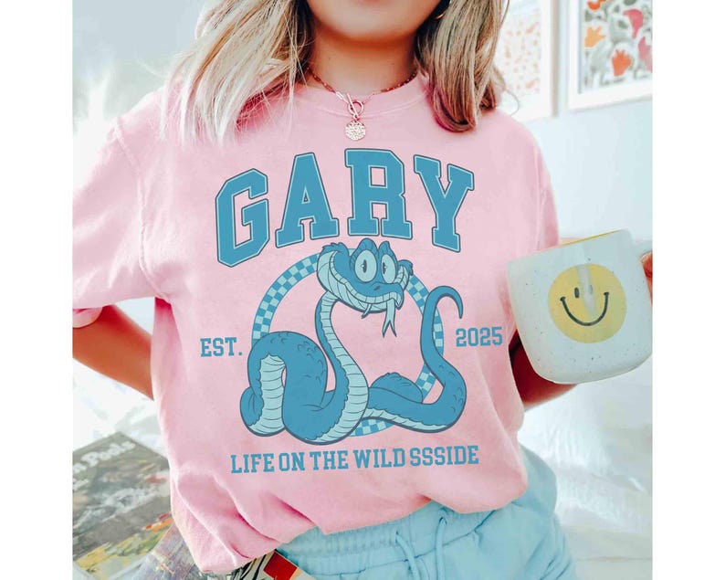 May include: Pink t-shirt with a blue cartoon snake graphic. The word "GARY" is arched across the top in blue block letters. Below the snake is the text "LIFE ON THE WILD SSSIDE" and the year "2025". A white mug with a yellow smiley face is held in the hand.
