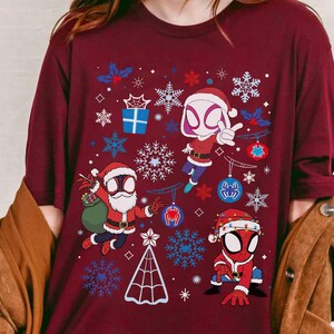 May include: Burgundy long-sleeve shirt featuring a festive design with Spider-Man characters in Santa Claus outfits. The graphic includes snowflakes, ornaments, and a Christmas tree. The shirt is a comfortable, casual wear.