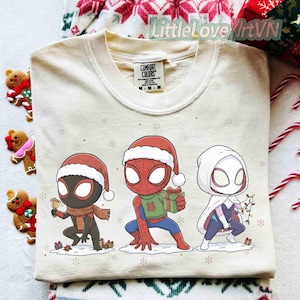 Może przedstawiać: Kremowa bluza Comfort Colors z trzema postaciami z kresk&oacute;wek Spider-Mana w czapkach Świętego Mikołaja. Jeden trzyma prezent, drugi dzwonek, a trzeci lampki choinkowe. Płatki śniegu i małe prezenty są rozrzucone.