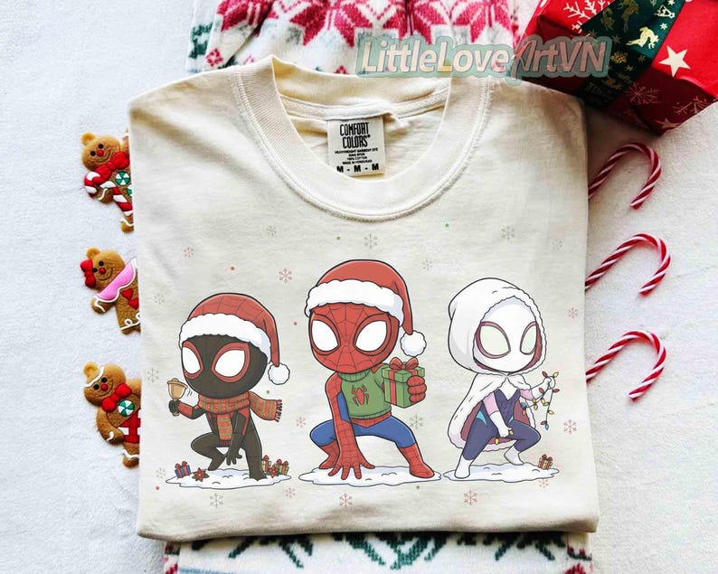 Może przedstawiać: Kremowa bluza Comfort Colors z trzema postaciami z kresk&oacute;wek Spider-Mana w czapkach Świętego Mikołaja. Jeden trzyma prezent, drugi dzwonek, a trzeci lampki choinkowe. Płatki śniegu i małe prezenty są rozrzucone.