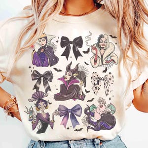 May include: Cream-colored t-shirt featuring illustrations of various Disney villains, including the Evil Queen, Maleficent, Cruella de Vil, and Ursula. The design incorporates bows and bats, creating a whimsical and thematic aesthetic.