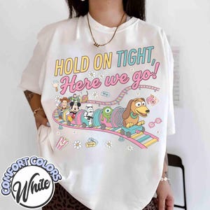 May include: White Comfort Colors t-shirt with a colorful graphic of a roller coaster featuring Disney characters and the text "HOLD ON TIGHT, Here we go!". The design includes Mickey Mouse, Woody, and Slinky Dog.