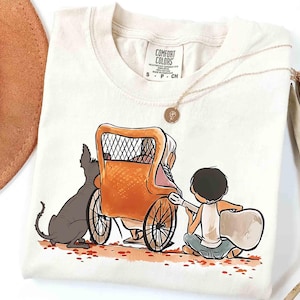 May include: Cream-colored t-shirt featuring a whimsical illustration of a person playing a guitar for another person in a carriage, with a dog. The shirt has a Comfort Colors tag. A gold necklace and a brown hat are also visible.