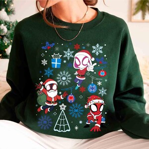 May include: Dark green sweatshirt featuring a festive design with cartoon Spider-Man characters in Santa hats. The graphic includes snowflakes, ornaments, and a Christmas tree. The design is set against a dark green background.