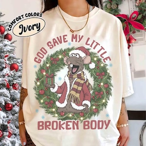 May include: Ivory-colored t-shirt with a Christmas-themed graphic. The graphic features a cartoon rat wearing a Santa hat and coat, surrounded by a wreath and the text "GOD SAVE MY LITTLE BROKEN BODY."