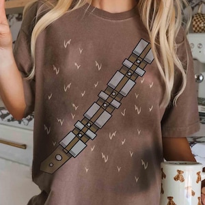 May include: Brown t-shirt with a bandolier graphic, featuring rectangular and circular details. The shirt has a crew neck and short sleeves. Small white "W" and "V" shapes are scattered across the shirt. A mug with bear illustrations is visible.