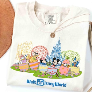 May include: Cream-colored t-shirt featuring Disney characters emerging from Easter eggs, with a castle and sphere in the background. The shirt has the words "Walt Disney World" printed at the bottom. A gold necklace and a brown hat are also in the image.