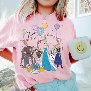 Może przedstawiać: R&oacute;żowa koszulka z kolorową grafiką postaci z kresk&oacute;wek, balon&oacute;w i tortu urodzinowego. Projekt zawiera cyfrę 5 i słowo "Name". Na zdjęciu trzymany jest biały kubek z uśmiechniętą buzią.