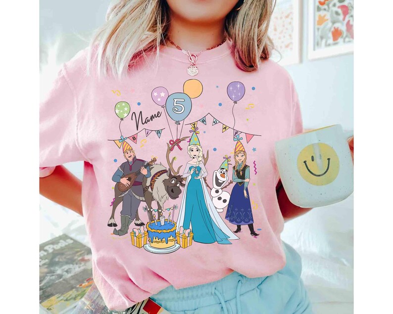 Może przedstawiać: R&oacute;żowa koszulka z kolorową grafiką postaci z kresk&oacute;wek, balon&oacute;w i tortu urodzinowego. Projekt zawiera cyfrę 5 i słowo "Name". Na zdjęciu trzymany jest biały kubek z uśmiechniętą buzią.