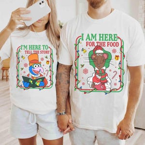 May include: Two white t-shirts with festive graphics. One displays Gonzo reading a book with the text "I'M HERE TO TELL THE STORY". The other shows a rat in a Santa hat and the text "I AM HERE FOR THE FOOD". Christmas-themed apparel.