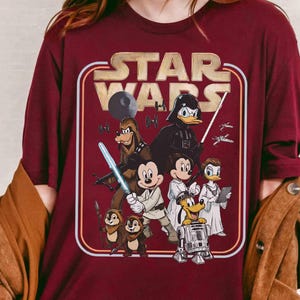 Puede incluir: Camiseta burdeos con el logo de Star Wars en un dise&ntilde;o retro. El gr&aacute;fico incluye varios personajes de Disney disfrazados de personajes de Star Wars, como Mickey Mouse como Luke Skywalker y Donald Duck como Darth Vader.