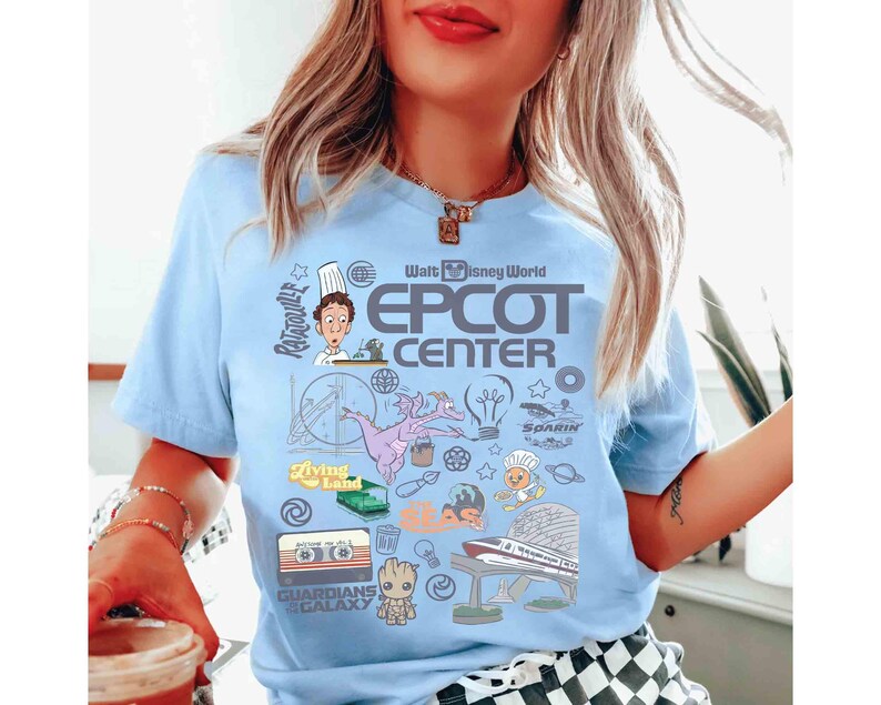Peut inclure: T-shirt bleu clair avec un motif graphique color&eacute;. Le motif comprend le texte "Walt Disney World Epcot Center" et des illustrations de personnages et d'ic&ocirc;nes du parc &agrave; th&egrave;me, comme R&eacute;my de Ratatouille et Figment.