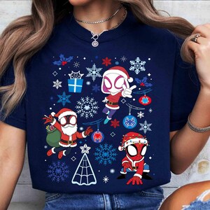May include: Navy blue t-shirt featuring a festive design with three Spider-Man characters in Santa hats, surrounded by snowflakes, ornaments, and a Christmas tree. The graphic is in red, white, and blue colors, perfect for the holiday season.