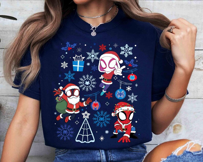 May include: Navy blue t-shirt featuring a festive design with three Spider-Man characters in Santa hats, surrounded by snowflakes, ornaments, and a Christmas tree. The graphic is in red, white, and blue colors, perfect for the holiday season.